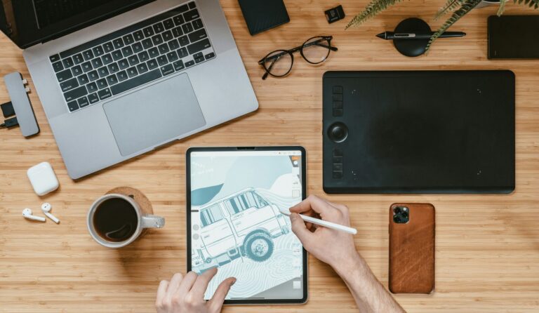Desk with a laptop, a pen tablet and an iPad showing a drawing in progress, with a stylus and coffee cup