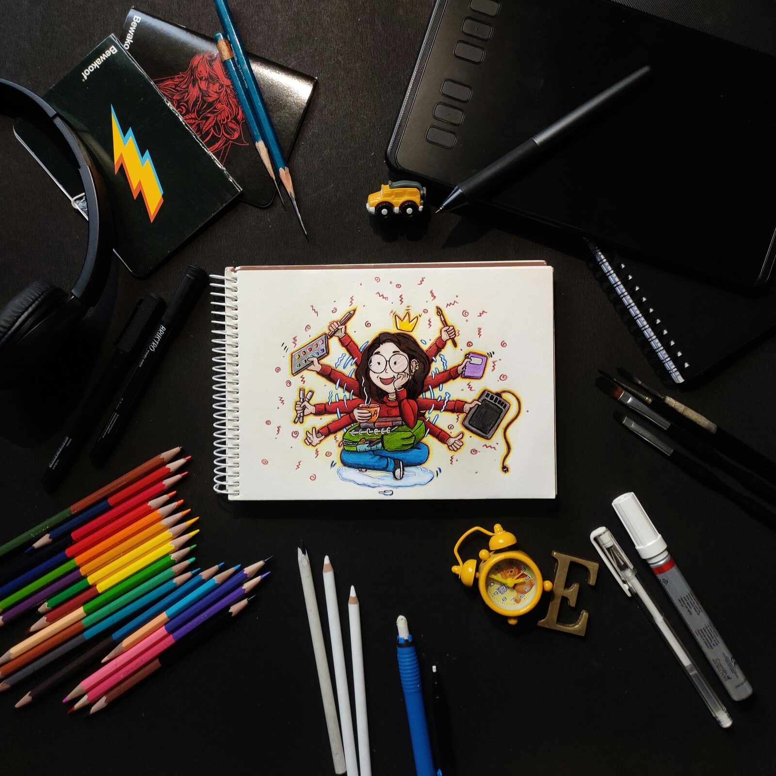 Flat lay of a graphics tablet and stylus beside a sketchbook, colored pencils and headphones